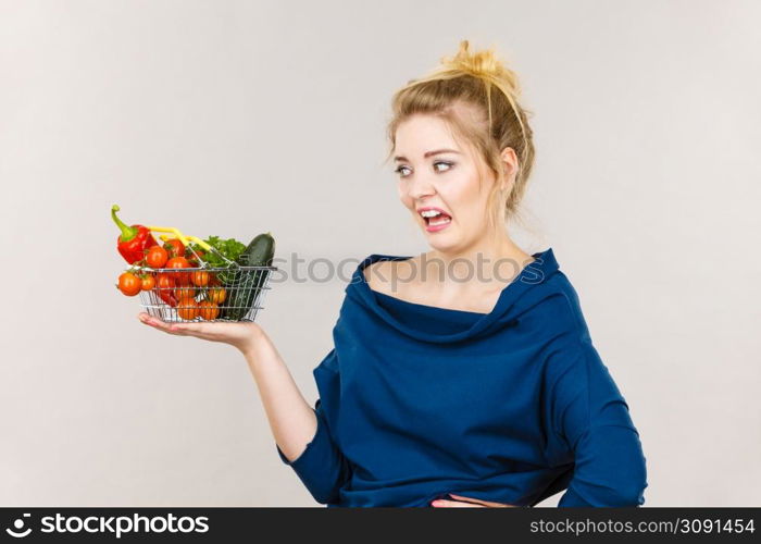 Adult woman do not like to eat vegetables, healthy food, vegetarian products. Female holding small shopping basket with green red vegetables, negative displeased face expression, on grey. Woman with vegetables, negative face expression