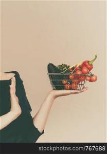 Adult woman do not like to eat vegetables, healthy food, vegetarian products. Female holding small shopping basket with green red vegetables, stop gesture, on grey. Woman with vegetables, stop gesture
