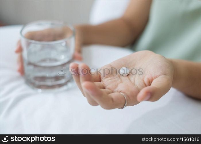 Adult woman holding pill and glass of water, taking medicine on bed in morning at home. Migraine, painkiller, headache, influenza, illness, sickness and healthcare concept