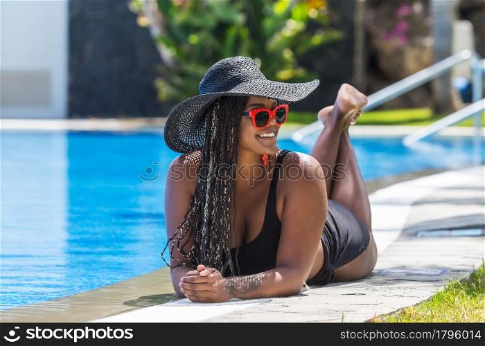 Adult woman in swimsuit lying on front on the edge of the pool looking to a side. African American woman with hat and sunglasses enjoying a summer day.. Adult woman in swimsuit lying on front on the edge of the pool looking to a side
