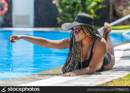 Adult woman in swimsuit lying on front on the edge of the pool touching the pool water. African American woman with hat and sunglasses enjoying a summer day.. Adult woman in swimsuit lying on front on the edge of the pool touching the pool water
