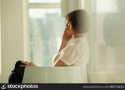 Adult woman sitting in chair and talking on phone against of window.. Side view of businesswoman talking on phone