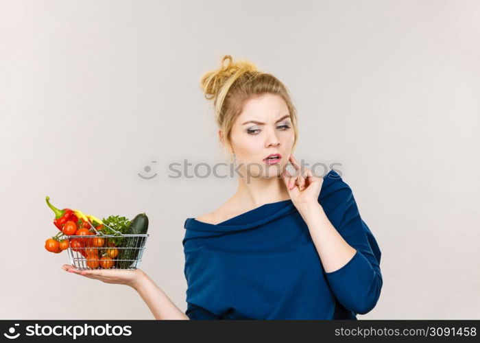 Adult woman with raw food, questioning healthy lifestyle recommendations, origin vegetagles. Female holding small shopping basket with products, thinking face expression. Woman with vegetables, thinking face expression