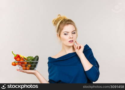 Adult woman with raw food, questioning healthy lifestyle recommendations, origin vegetagles. Female holding small shopping basket with products, thinking face expression. Woman with vegetables, thinking face expression