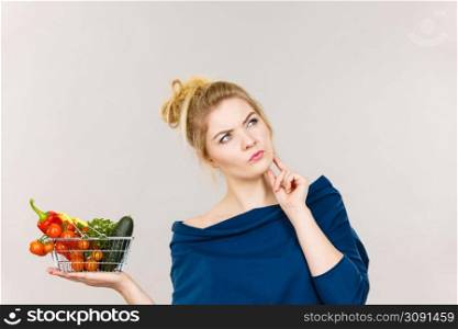 Adult woman with raw food, questioning healthy lifestyle recommendations, origin vegetagles. Female holding small shopping basket with products, thinking face expression. Woman with vegetables, thinking face expression