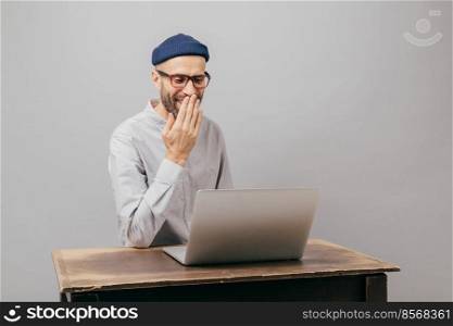 Adult young male blogger chats with followers, sends feedback, enjoys spending free time, smiles as reads funny news, covers mouth with palm, wears formal clothes, isolated over grey background