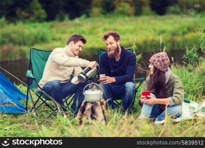 adventure, travel, tourism and people concept - group of smiling friends cooking food in dixie sitting around bonfire outdoors