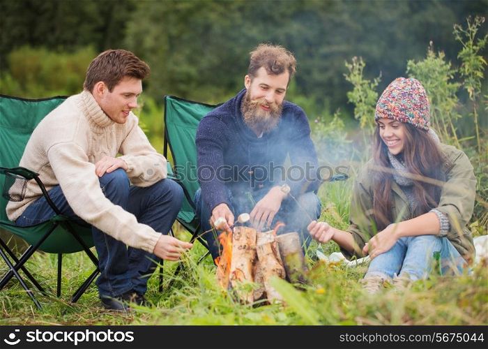 adventure, travel, tourism and people concept - group of smiling friends sitting on chairs around bonfire in camping