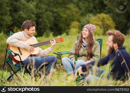adventure, travel, tourism and people concept - group of smiling friends with guitar sitting on chairs in camping
