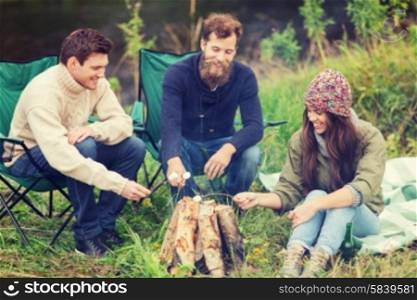 adventure, travel, tourism and people concept - group of smiling friends with marshmallow sitting around bonfire in camping