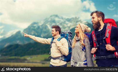 adventure, travel, tourism, hike and people concept - group of smiling friends with backpacks pointing finger over alpine mountains background