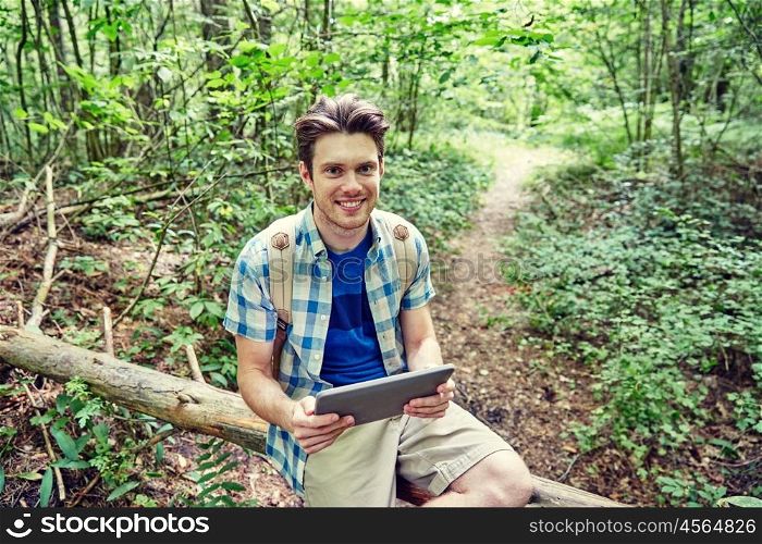 adventure, travel, tourism, hike and people concept - happy young man with backpack and tablet pc computer sitting on fallen tree trunk in woods