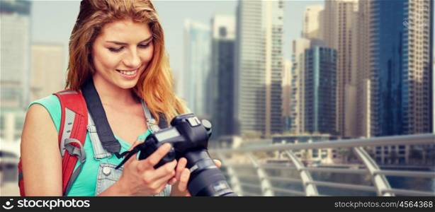 adventure, travel, tourism, hike and people concept - happy young woman with backpack and camera photographing over dubai city waterfront background