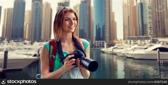 adventure, travel, tourism, hike and people concept - happy young woman with backpack and camera photographing over dubai city and harbor with boats background