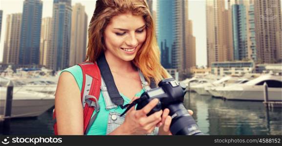 adventure, travel, tourism, hike and people concept - happy young woman with backpack and camera photographing over dubai city and harbor with boats background