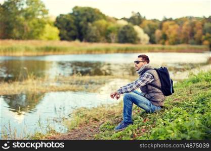 adventure, travel, tourism, hike and people concept - man in sunglasses with backpack resting on river bank