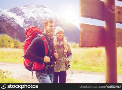 adventure, travel, tourism, hike and people concept - smiling couple with backpacks standing at signpost over alpine mountains and hills background