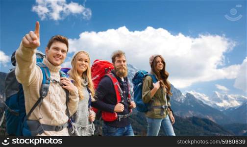 adventure, travel, tourism, hike and people concept - smiling friends walking with backpacks and pointing finger to something over mountains background