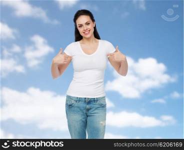 advertisement, clothing and people concept - happy smiling young woman or teenage girl in white t-shirt pointing finger to herself over blue sky and clouds background