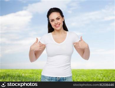 advertisement, clothing and people concept - happy smiling young woman or teenage girl in white t-shirt pointing finger to herself over blue sky and grass background