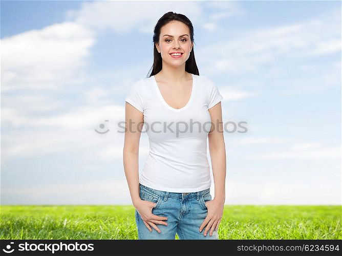 advertisement, clothing and people concept - happy smiling young woman or teenage girl in white t-shirt over blue sky and grass background