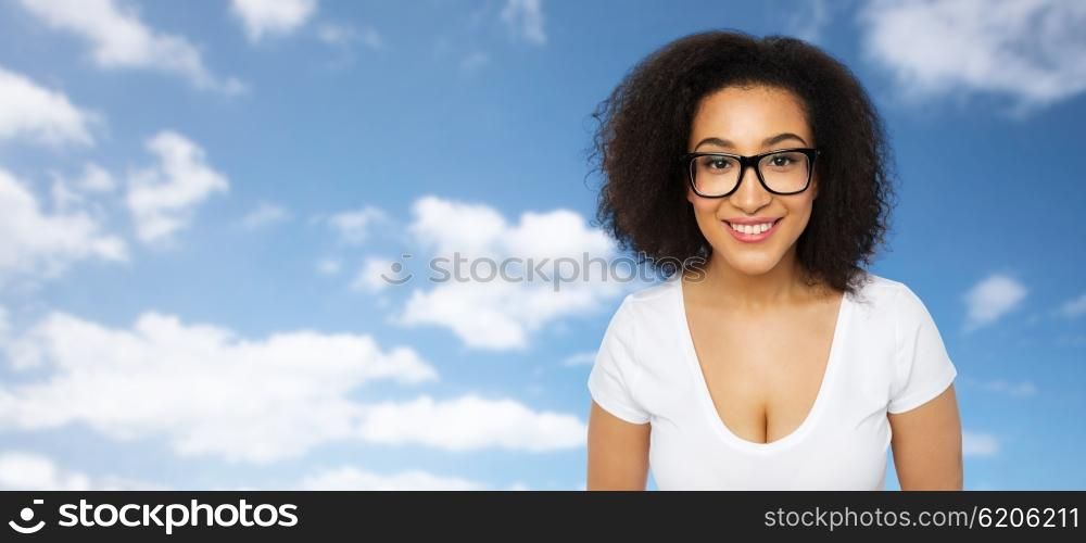 advertisement, education, ethnicity, vision and people concept - happy smiling young african woman or teenage student girl in eyeglasses and white t-shirt over blue sky and clouds background