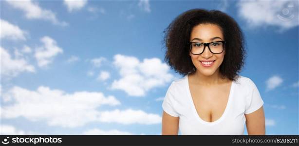 advertisement, education, ethnicity, vision and people concept - happy smiling young african woman or teenage student girl in eyeglasses and white t-shirt over blue sky and clouds background