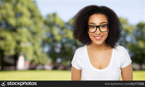 advertisement, education, ethnicity, vision and people concept - happy smiling young african woman or teenage student girl in eyeglasses and white t-shirt over summer park background