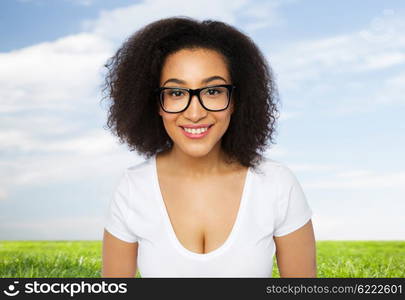 advertisement, education, ethnicity, vision and people concept - happy smiling young african woman or teenage student girl in eyeglasses and white t-shirt over blue sky and grass background