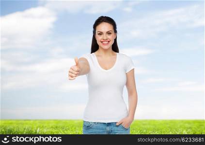 advertisement, gesture, clothing and people concept - happy smiling young woman or teenage girl in white t-shirt showing thumbs up over blue sky and grass background