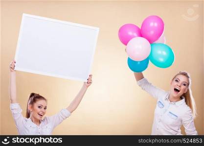Advertisement, holidays celebration concept. Smiling joyful girls holding presentation board, banner sign and colorful balloons, having fun. Two girls with blank board and balloons