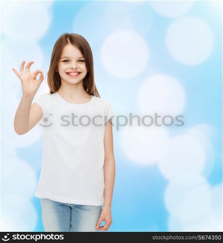 advertising, childhood and people - smiling little girl in white blank t-shirt showing ok gesture over blue background