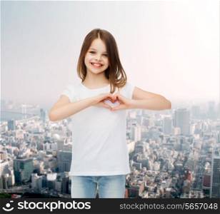 advertising, childhood, charity and people concept - smiling little girl in white t-shirt making heart-shape gesture over city background