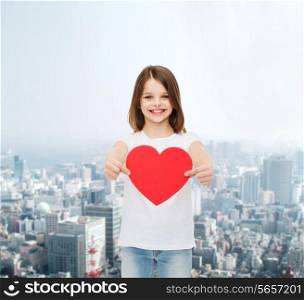 advertising, childhood, charity and people concept - smiling little girl in white t-shirt holding red heart cutout over city background