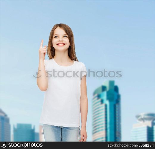 advertising, childhood, city, gesture and people concept - smiling girl in white t-shirt pointing finger up over business center background