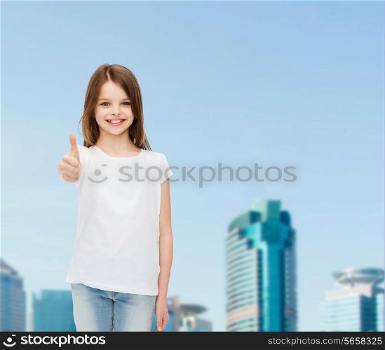 advertising, childhood, city, gesture and people concept - smiling girl in white t-shirt showing thumbs up over business center background