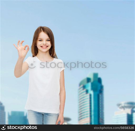 advertising, childhood, city, gesture and people concept - smiling girl in white t-shirt showing ok sign over business center background