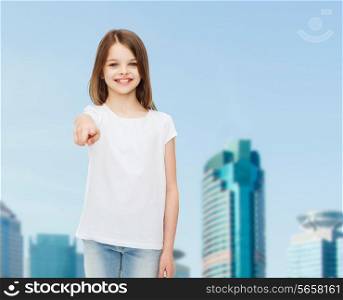 advertising, childhood, gesture and people concept - smiling girl in white t-shirt pointing finger on you over business center background