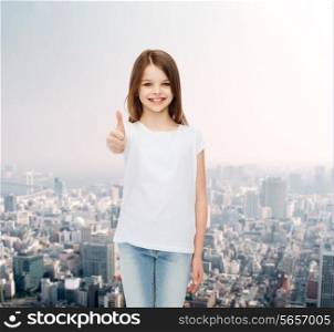 advertising, childhood, gesture and people concept - smiling little girl in white blank t-shirt showing thumbs up over city background