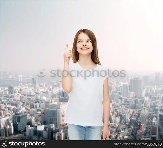 advertising, childhood, gesture and people concept - smiling little girl in white blank t-shirt pointing her finger up over city background