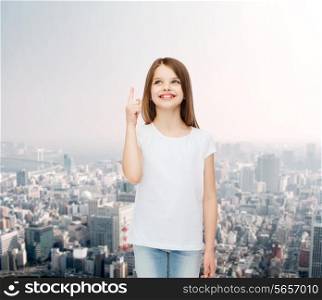 advertising, childhood, gesture and people concept - smiling little girl in white blank t-shirt pointing her finger up over city background