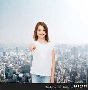 advertising, childhood, gesture and people concept - smiling little girl in white blank t-shirt pointing finger on you over city background