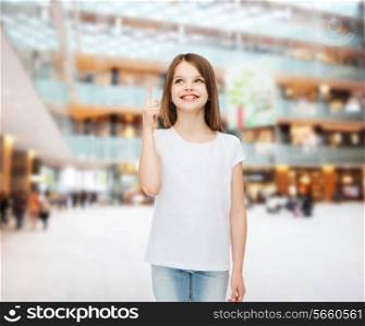 advertising, childhood, gesture, consumerism and people - smiling girl in white t-shirt pointing finger up over shopping center background