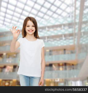 advertising, childhood, gesture, consumerism and people - smiling little girl in white blank t-shirt showing ok sign over shopping center background