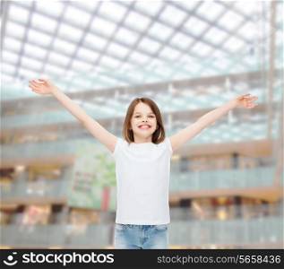advertising, childhood, gesture, consumerism and people - smiling little girl in white blank t-shirt with stretched out arms over shopping center background