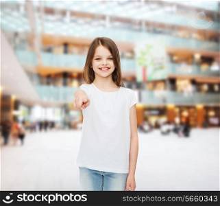 advertising, childhood, gesture, consumerism - smiling girl in white t-shirt pointing finger on you over shopping center background