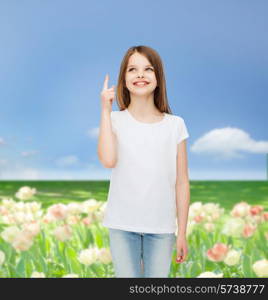 advertising, childhood, nature, gesture and people concept - smiling girl in white t-shirt pointing finger up over flower field background