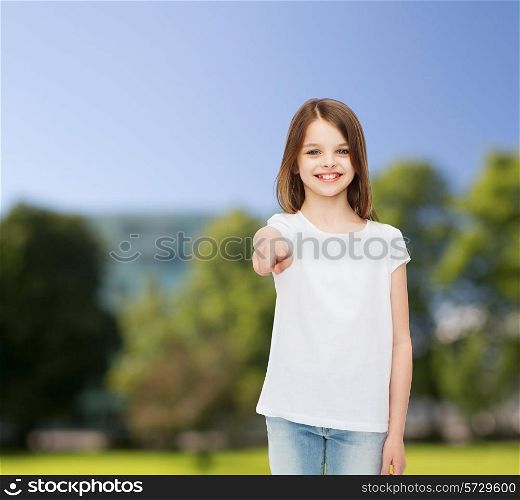 advertising, childhood, nature, gesture and people - smiling little girl in white blank t-shirt pointing finger on you over green park background
