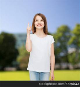 advertising, childhood, nature, gesture and people - smiling little girl in white t-shirt pointing her finger up over green park background