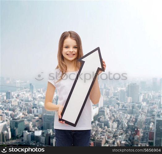 advertising, direction and childhood concept - smiling little girl with white blank arrow pointing up city background